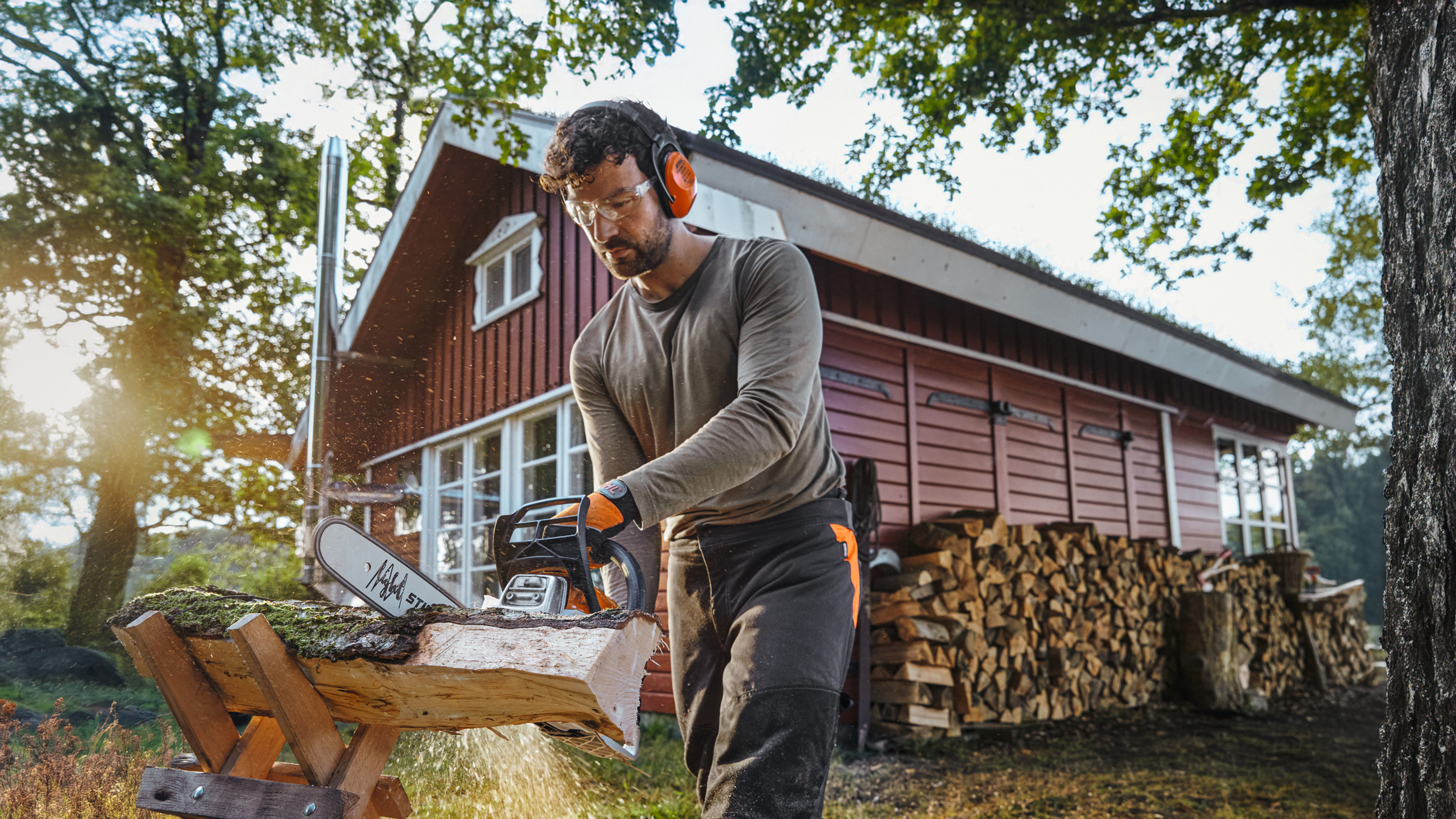 Motosierra STIHL con la que un hombre corta un tronco delante de una cabaña de leña roja con una pila de leña