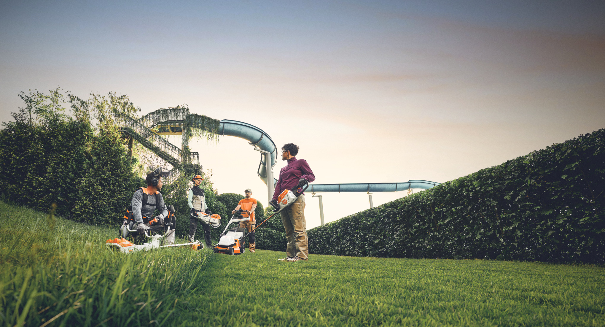 Un grupo de personas trabaja con máquinas de jardín STIHL en un jardín, con un gran seto y toboganes de agua al fondo. Están equipados con desbrozadoras, cortacéspedes y equipos de protección.