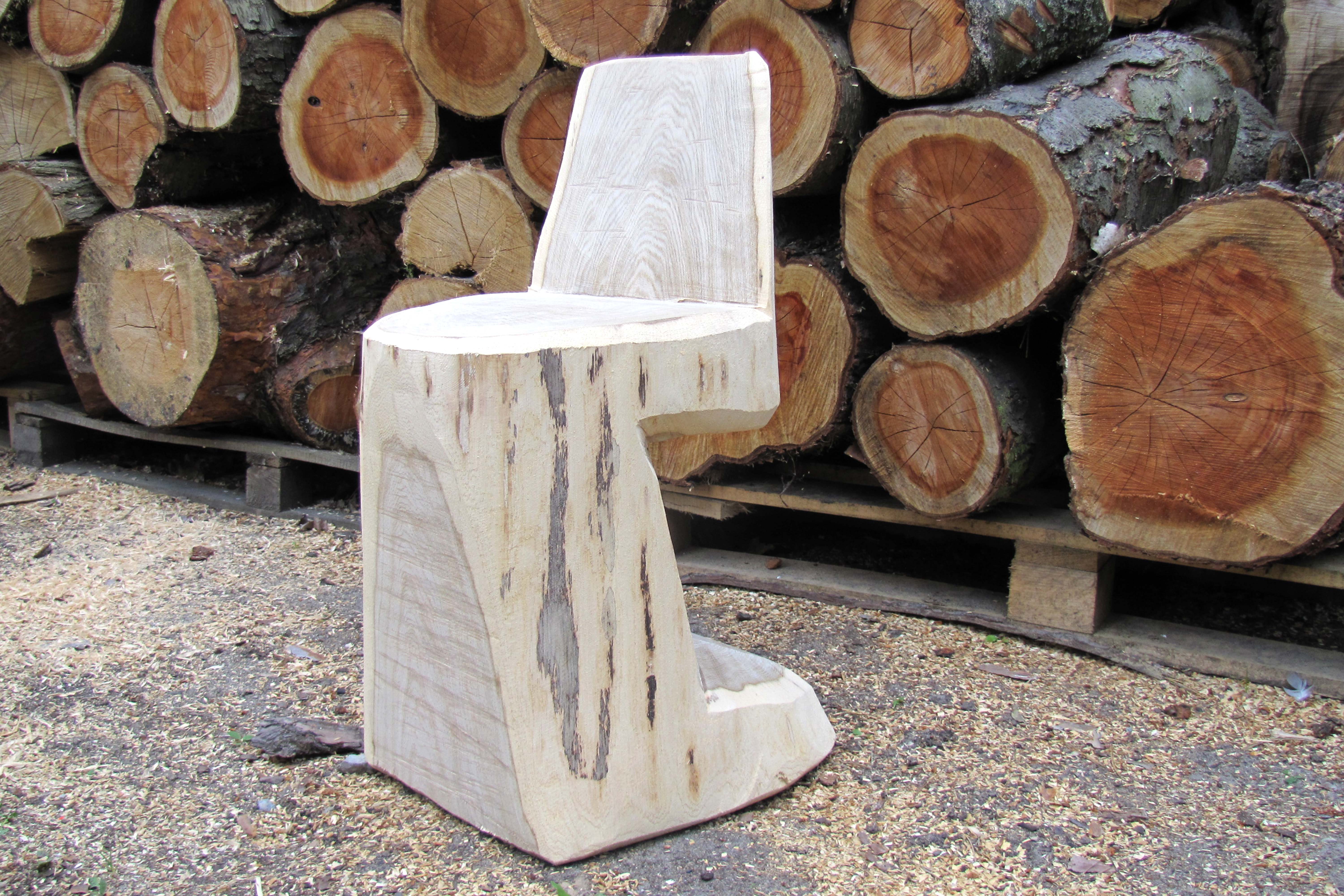 Una silla de madera artesanal construida a partir de un tronco delante de troncos de árboles.