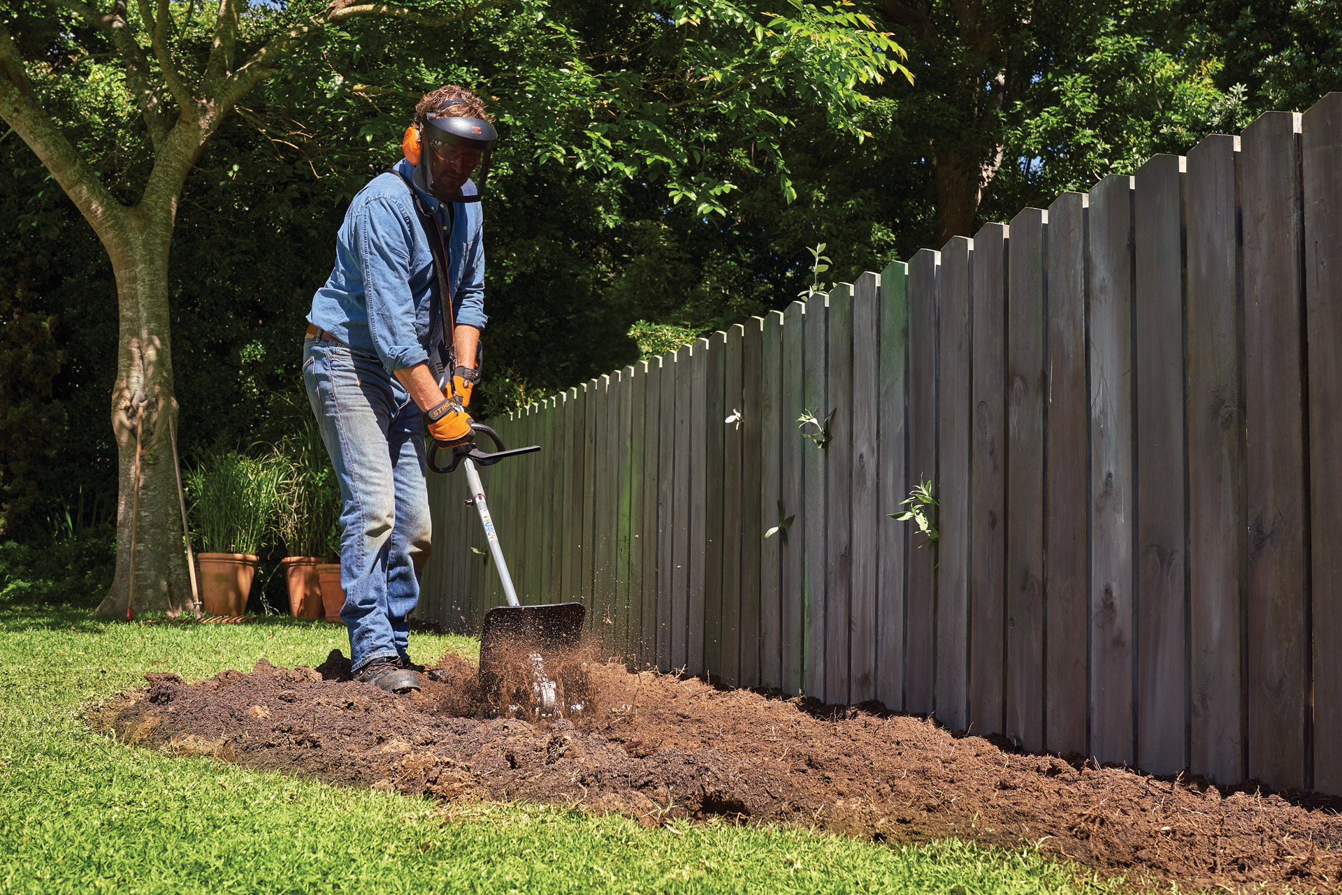 Una persona con equipo de protección cultiva la tierra en un jardín utilizando un STIHL KombiSystem con fresadora.