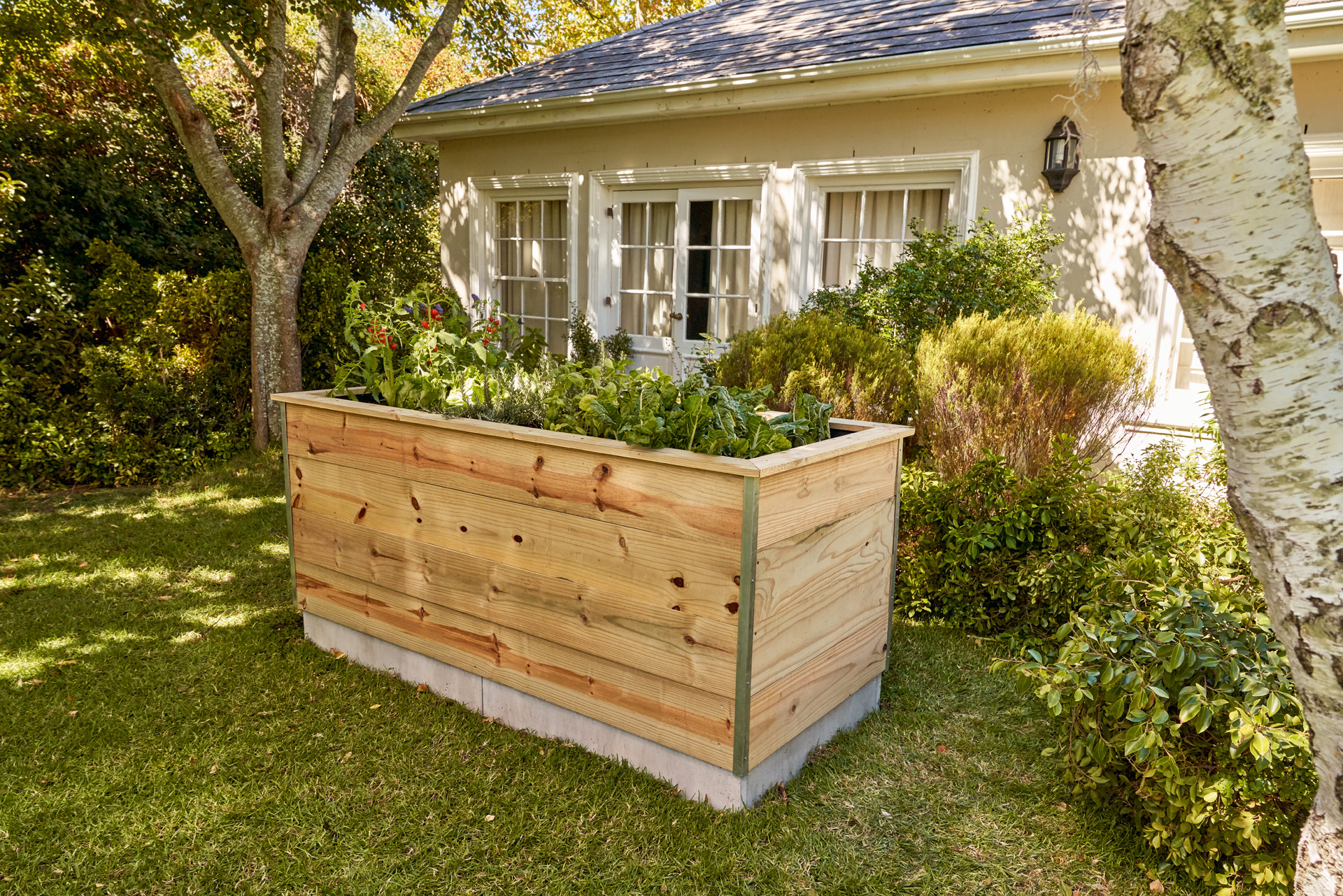 Una mesa de cultivo para huertos urbanos hecha con tablas de madera, en un jardín frente a una casa.