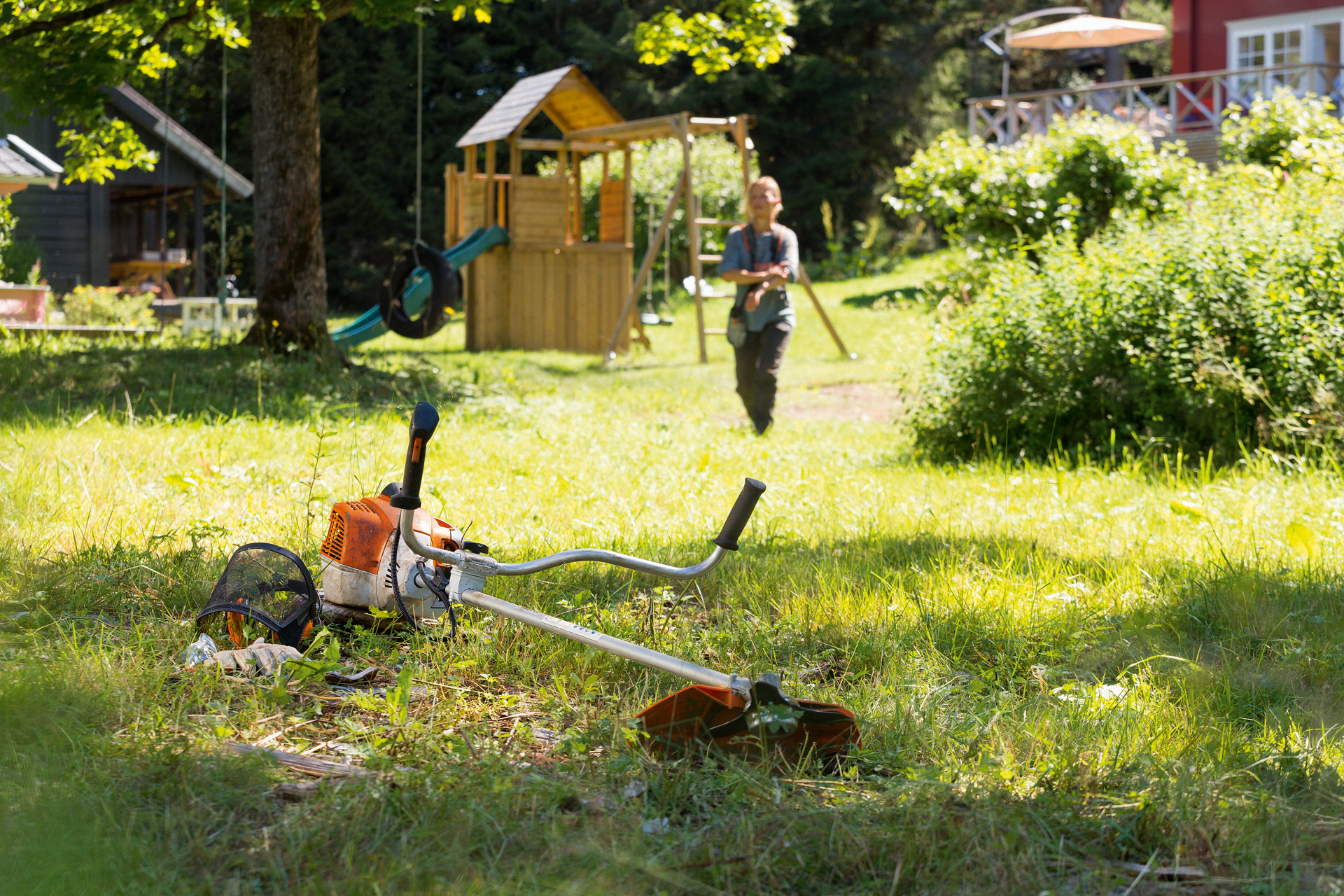 Una desbrozadora STIHL FS 240 en el suelo en un jardín con columpios y un hombre acercándose a la máquina.