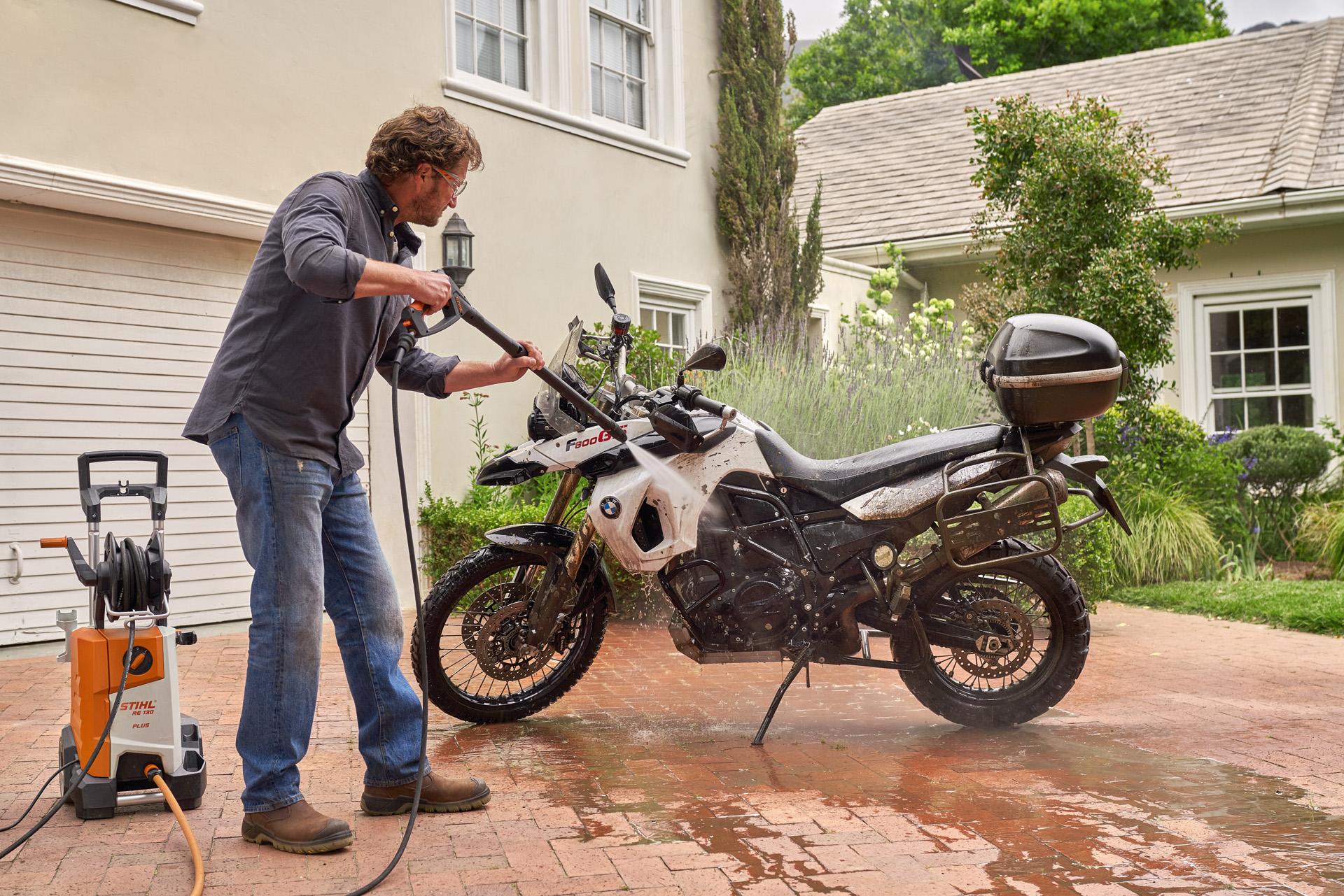 Un hombre utiliza una hidrolimpiadora STIHL RE 130 Plus para lavar una moto con agua a presión en un garaje de una casa.