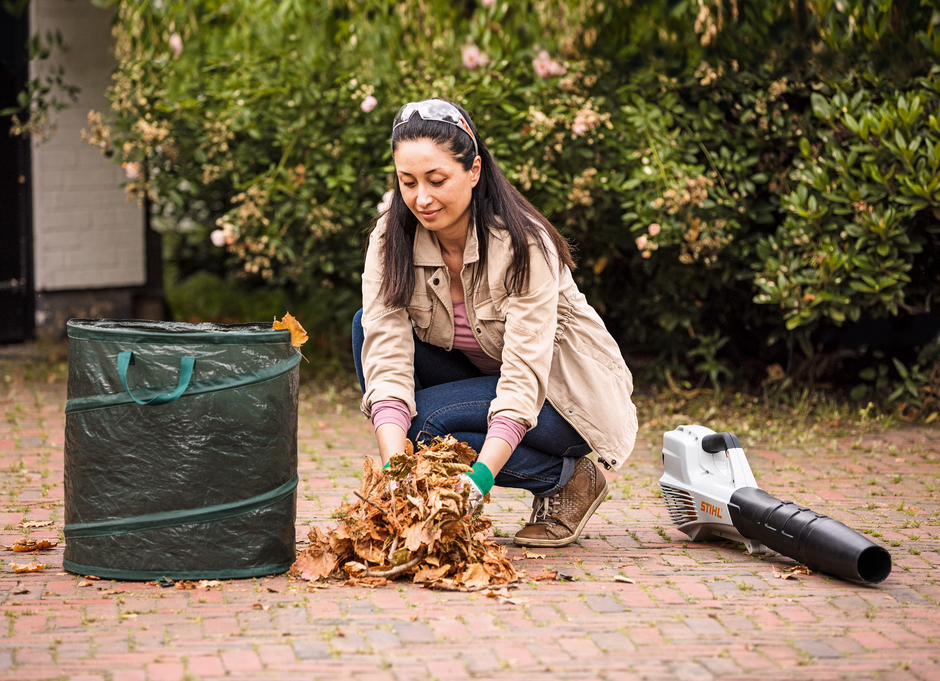 Una mujer ocupada en la tarea de recoger hojas secas de su jardín, junto a ella en el suelo un soplador STIHL BGA 56