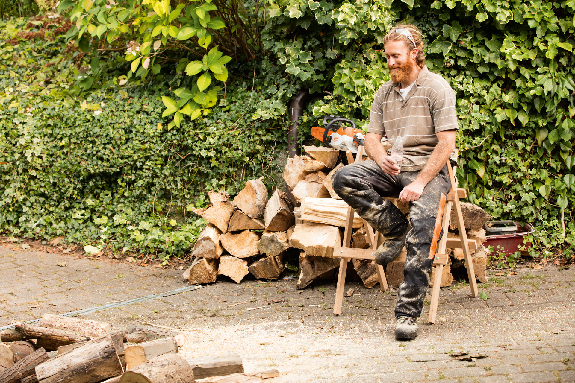 Hombre sentado en un banco para serrar delante de una pila de troncos partidos.