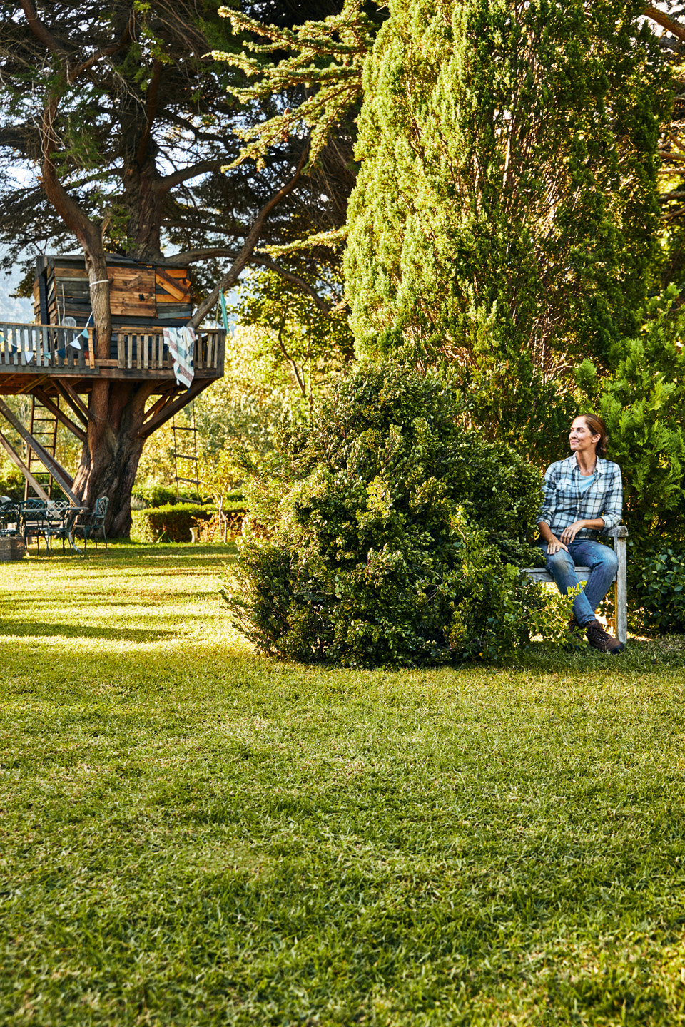Una mujer está sentada en un banco en un jardín con arbustos y césped, con una casa de árbol en el fondo